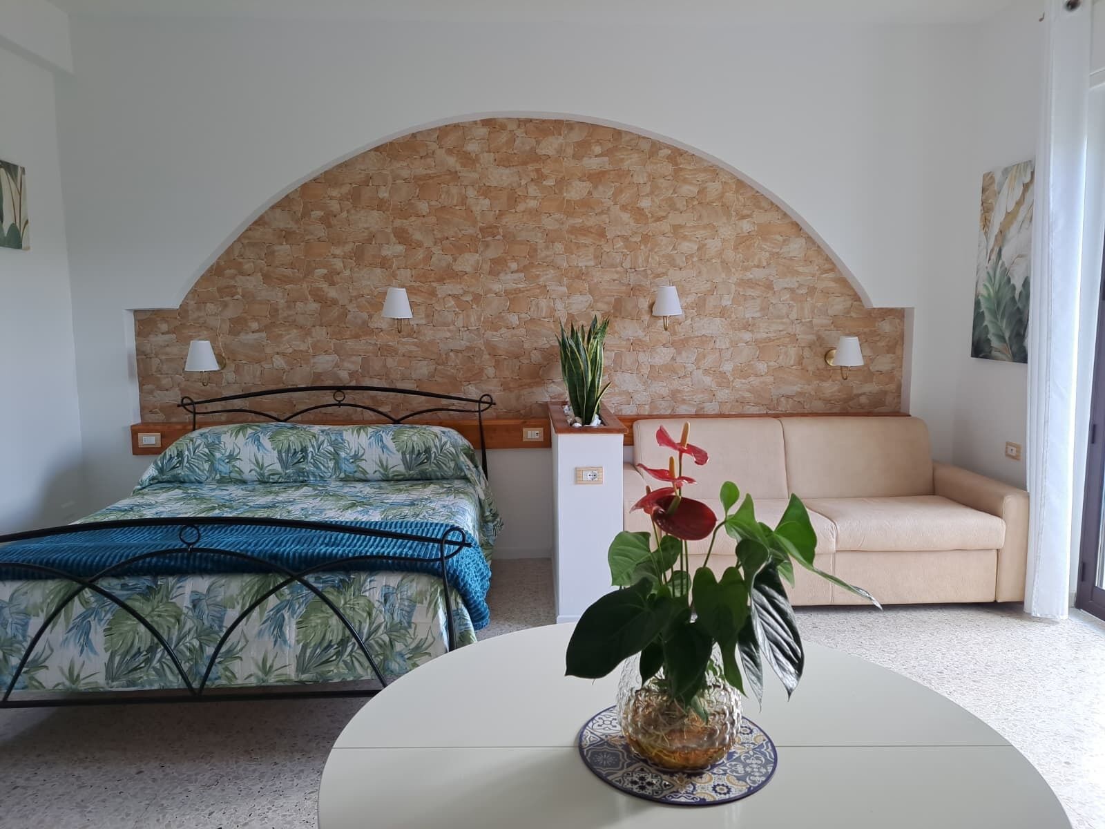 A bedroom with a bed, a sofa, a table with a plant, and a stone wall.