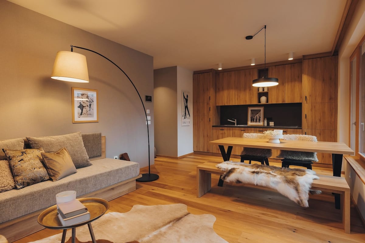 A modern living room with a sofa, a coffee table, a dining table with benches, and a kitchen area with wooden cabinets and a TV.
