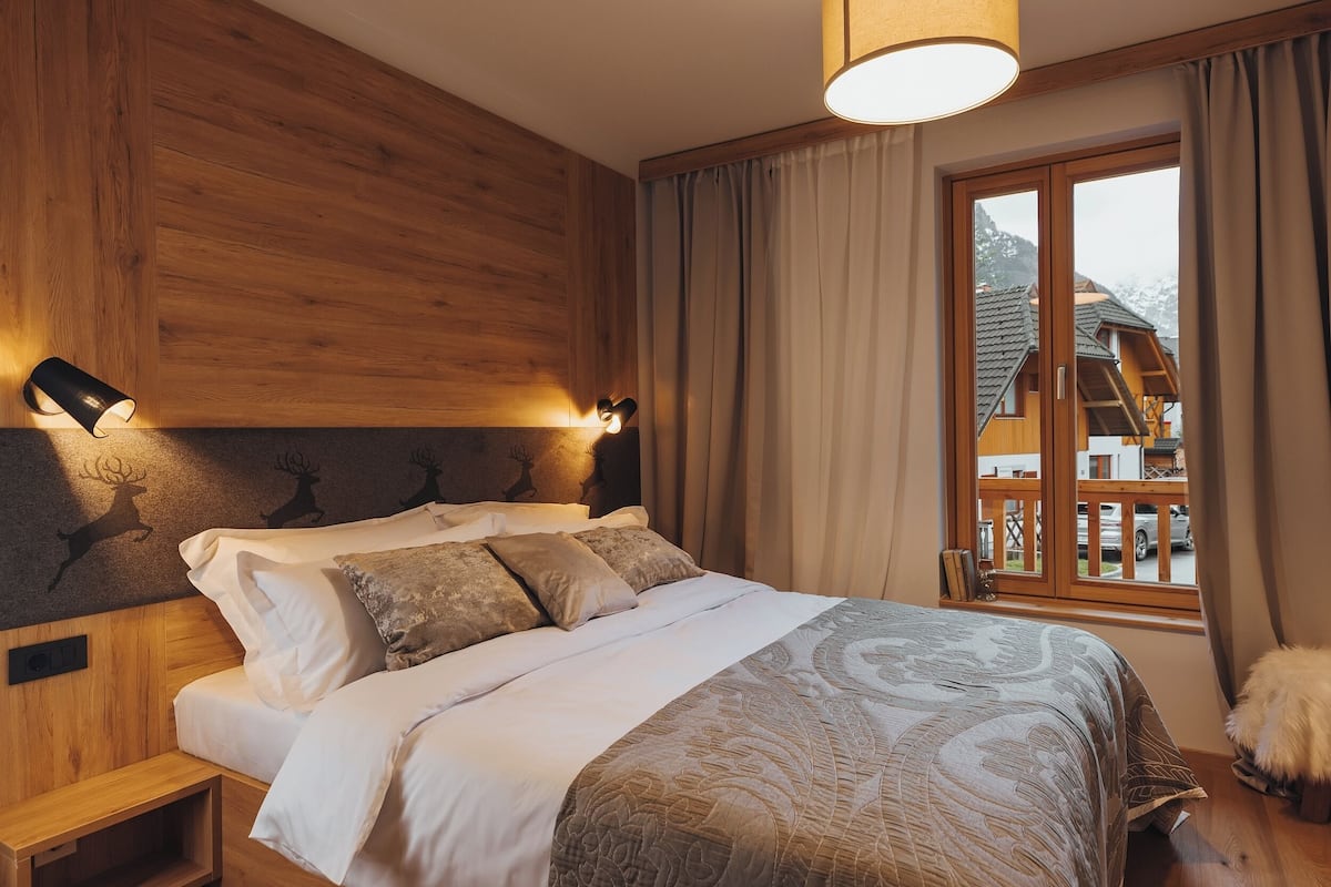 A bedroom with a wooden headboard, a large bed with white and patterned bedding, a nightstand, and a window with curtains.