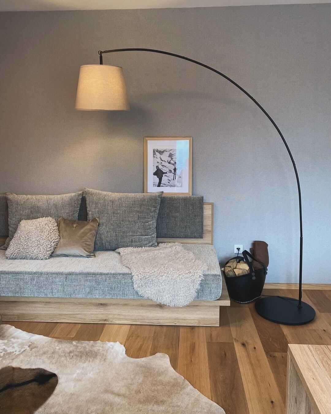 Ein modernes Wohnzimmer mit grauem Sofa, einer schwarzen Stehlampe, einem gerahmten Bild an der Wand und einem schwarzen Korb mit Holzscheiten.