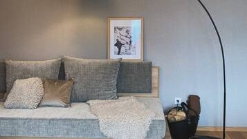 Ein modernes Wohnzimmer mit grauem Sofa, einer schwarzen Stehlampe, einem gerahmten Bild an der Wand und einem schwarzen Korb mit Holzscheiten.