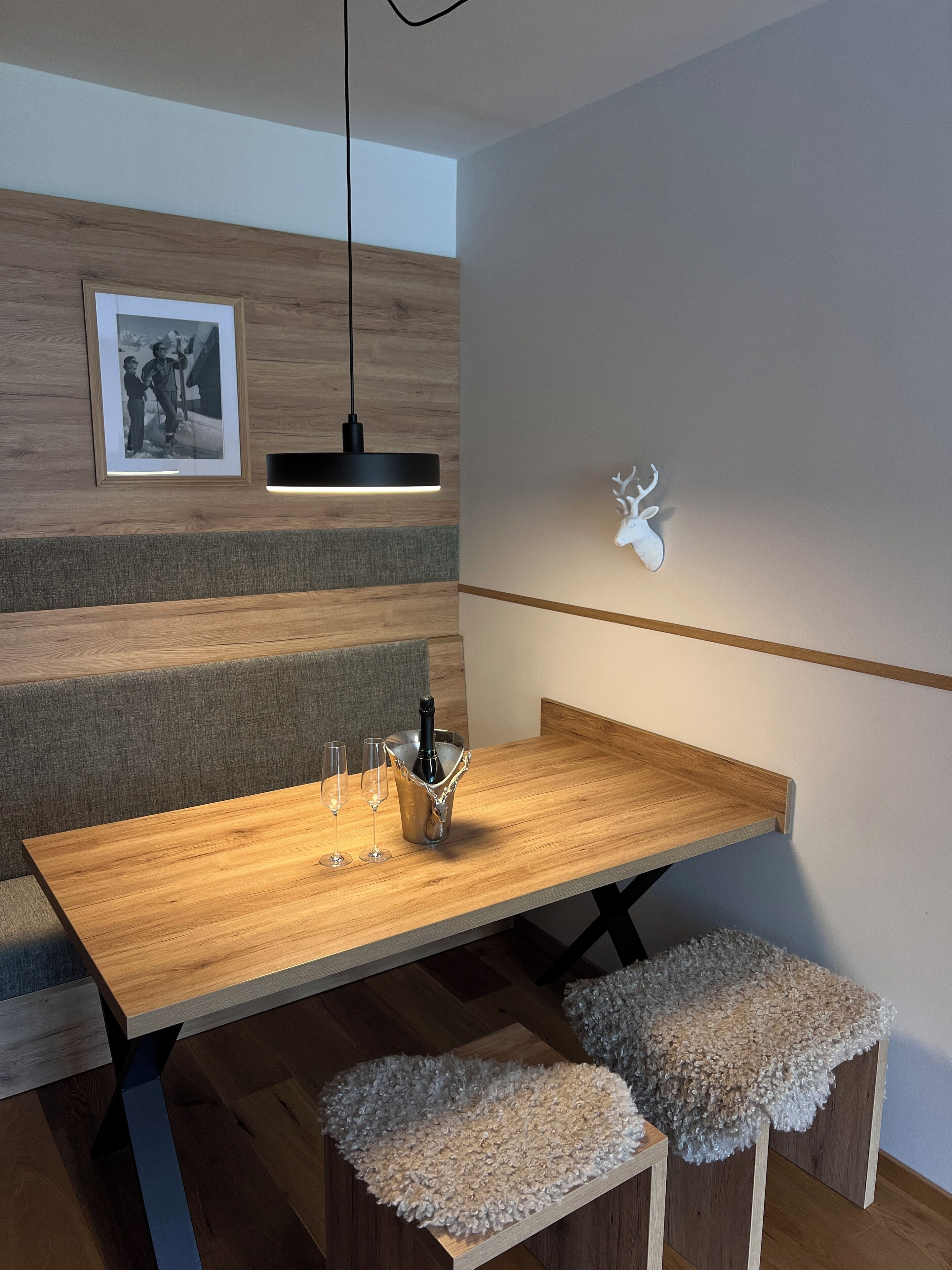 A modern dining area with a wooden table, a framed photograph, a pendant light, and a mounted deer head.
