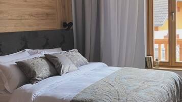 Un dormitorio con cabecera de madera, cama con ropa de cama blanca y un plaid estampado, una ventana con cortinas y vistas a edificios.