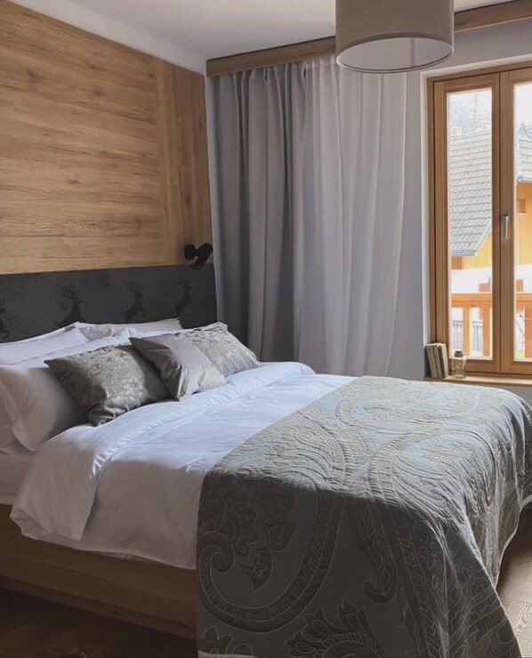 A bedroom with a wooden headboard, a bed with white bedding and patterned throw, a window with curtains, and a view of buildings outside. - Goldhorn Chalet (Kranjska Gora)