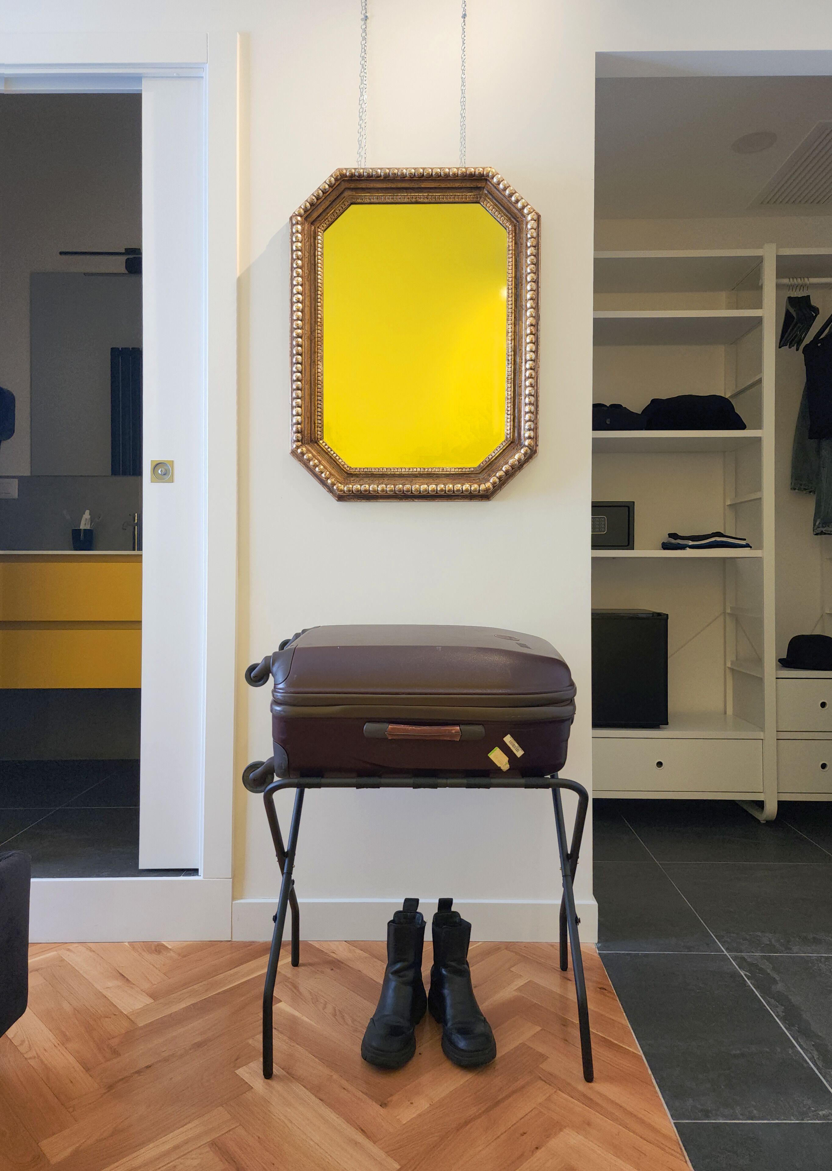 A room with a wooden floor, a suitcase on a stand, a pair of boots, and a mirror with a decorative frame.