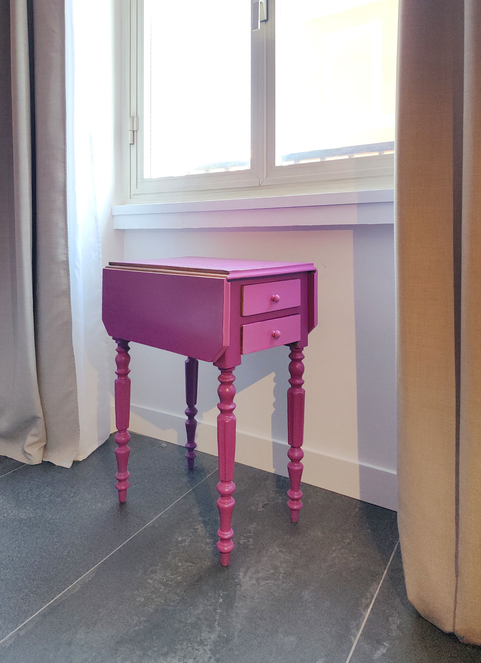 Una scrivania di legno rosa con due cassetti, posta di fronte a una finestra con persiane bianche.
