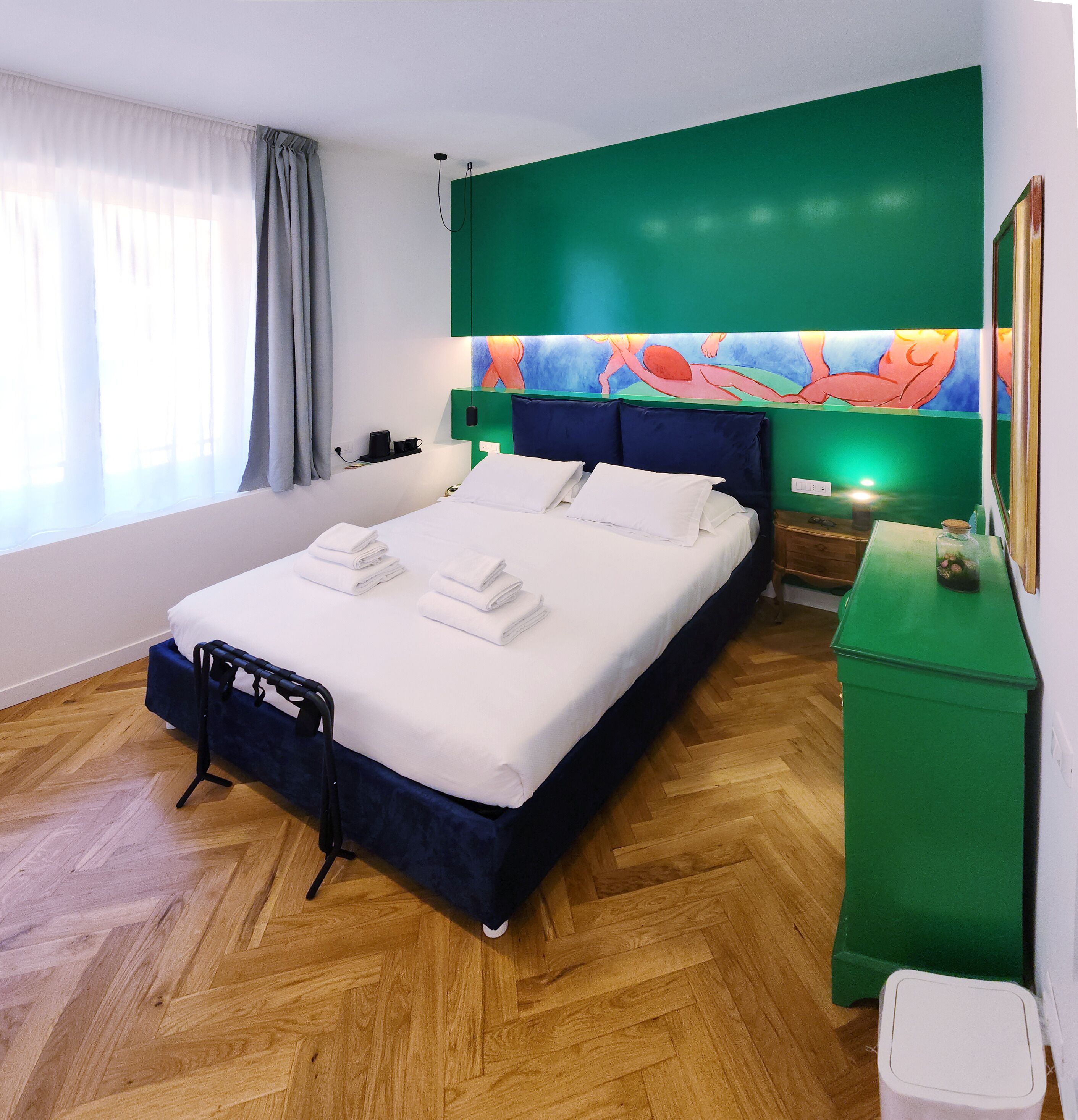 A hotel room with a bed, a green nightstand, a wooden floor, and a window with curtains.