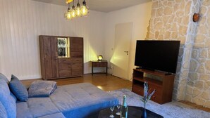 A living room with a sectional sofa, a flat-screen TV, a coffee table with drinks, and a stone wall.