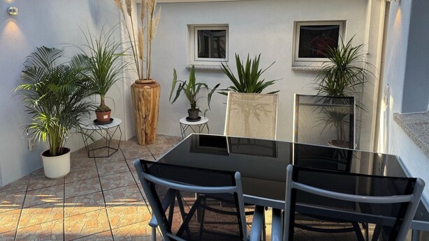 A patio with a glass table and chairs, surrounded by potted plants and a tiled floor.