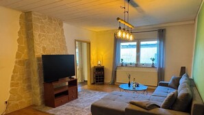 A living room with a stone wall, a large TV on a wooden stand, a grey sofa, a coffee table, and a hanging light fixture.