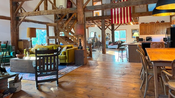 En rummelig loftrum med synlige træbjælker, et stort amerikansk flag og et hyggeligt siddeområde med sofa og stole.