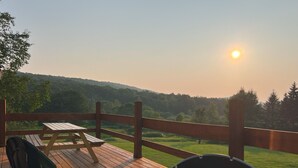En træterrasse med to Adirondack-stole og et picnicbord, med udsigt over et græslandskab og bakker.