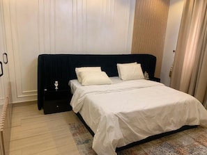 A neatly made bed with white linens and a dark headboard in a room with a wooden floor and a patterned rug.
