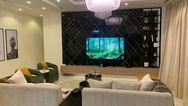 Un salon moderne doté d’un grand téléviseur à écran plat, de canapés verts et blancs, d’un lustre et d’un mur noir orné d’un motif en losanges.