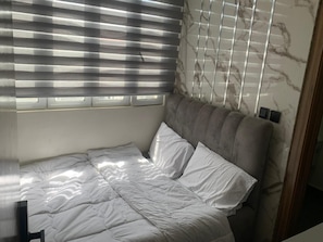 A bed with a grey upholstered headboard, white bedding, and a window with horizontal blinds.