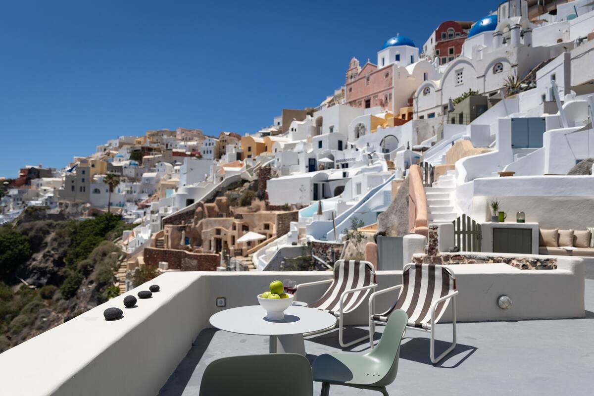 A terrace with a table and chairs, overlooking a hillside of white buildings with blue domes.