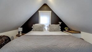 Ein Schlafzimmer mit schrägen Wänden, einem Bett mit gesteppter Bettdecke, zwei Nachttischen mit Lampen und einem Fenster mit Jalousien.