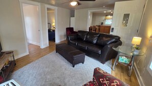 A living room with a leather sofa, ottoman, and a glass coffee table. There is a TV on a wooden stand and a small table with a lamp. The room has wooden flooring and white walls.