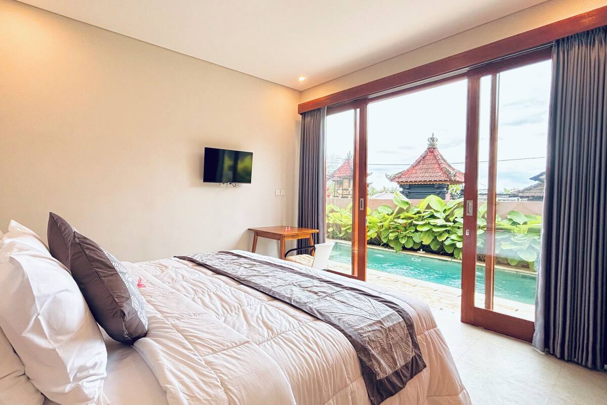 A bedroom with a bed, a TV, a small table, and a view of a pool and traditional buildings.