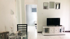 A living room with a TV on a white cabinet, a dining table with chairs, and a kitchen area in the background. - Beach and Bliss E32 - Ocho Rios Retreats (Ocho Rios)
