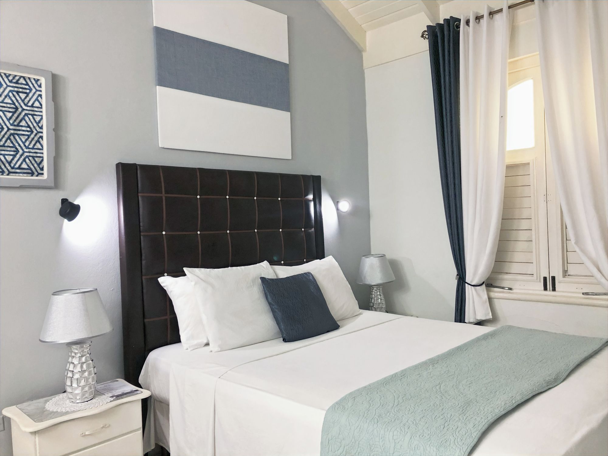 A hotel room with a bed, two bedside lamps, a nightstand, a decorative pillow, a wall art piece, and a window with curtains.