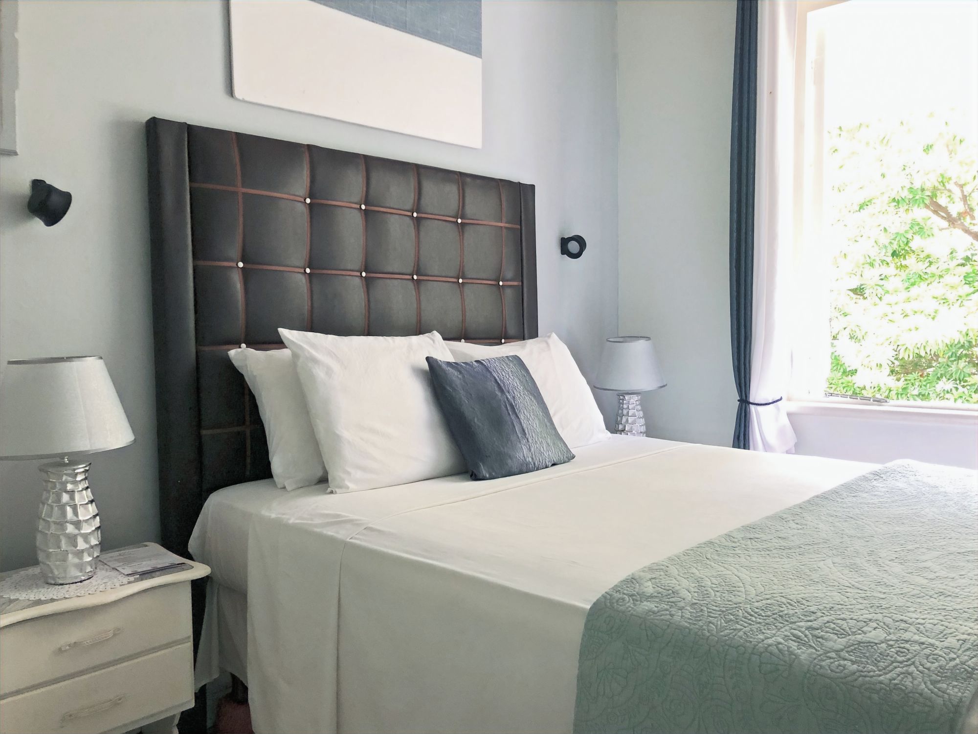 A hotel room with a large bed, two bedside lamps, a nightstand, and a window with a view of greenery.