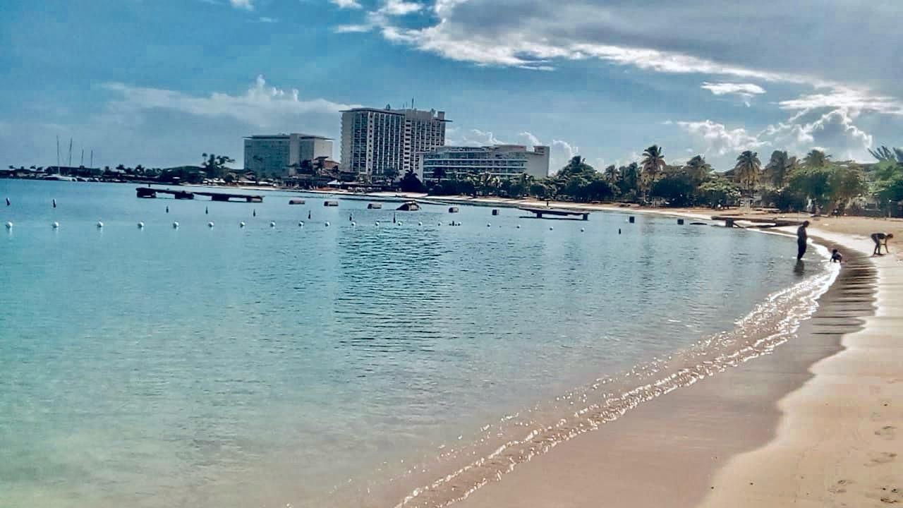 Une plage avec une eau cristalline, un rivage sablonneux et des bâtiments en arrière-plan.
