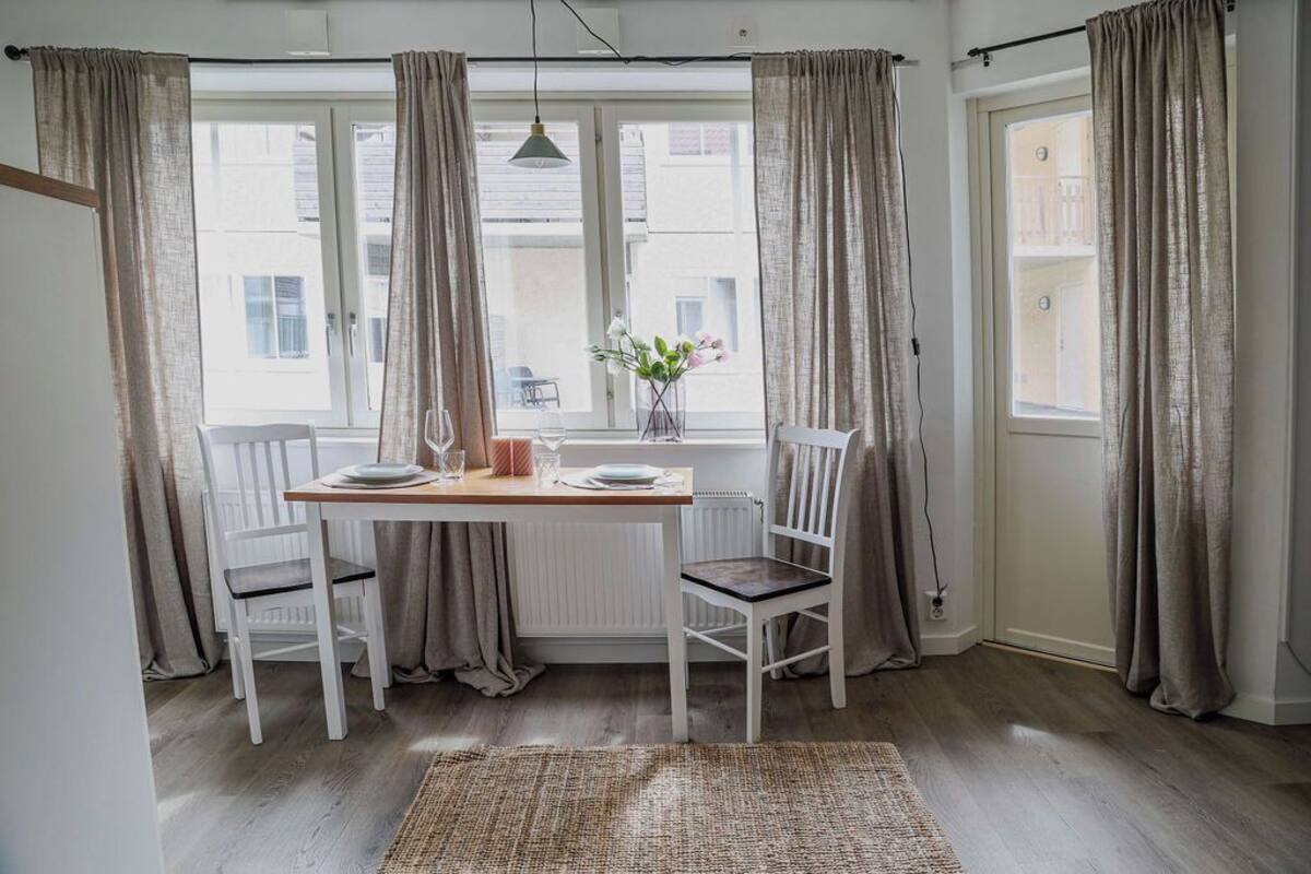 Ein Essbereich mit Tisch für zwei Personen, weißen Stühlen, einer Blumenvase und Blick durch das Fenster.
