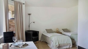 A hotel room with a dining table set for two, a black armchair, two single beds with pillows, a floor lamp, and a view of a wooden staircase. - GotlandStay Volontären (Visby)