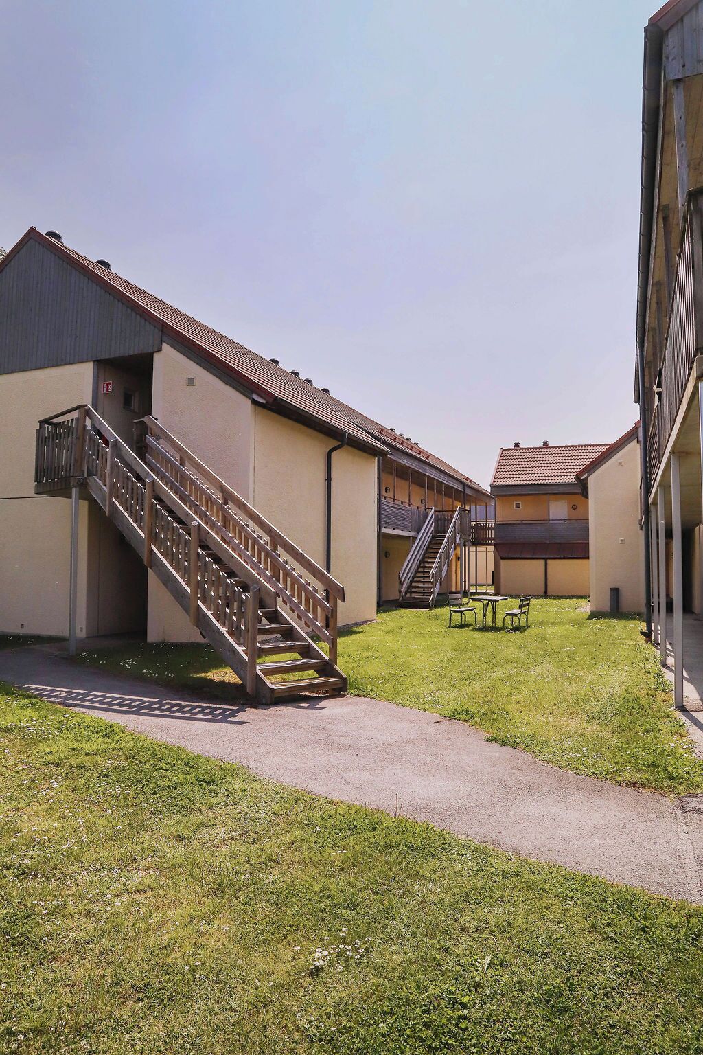 A building with a staircase leading to a covered area, a grassy courtyard, and a small seating area.