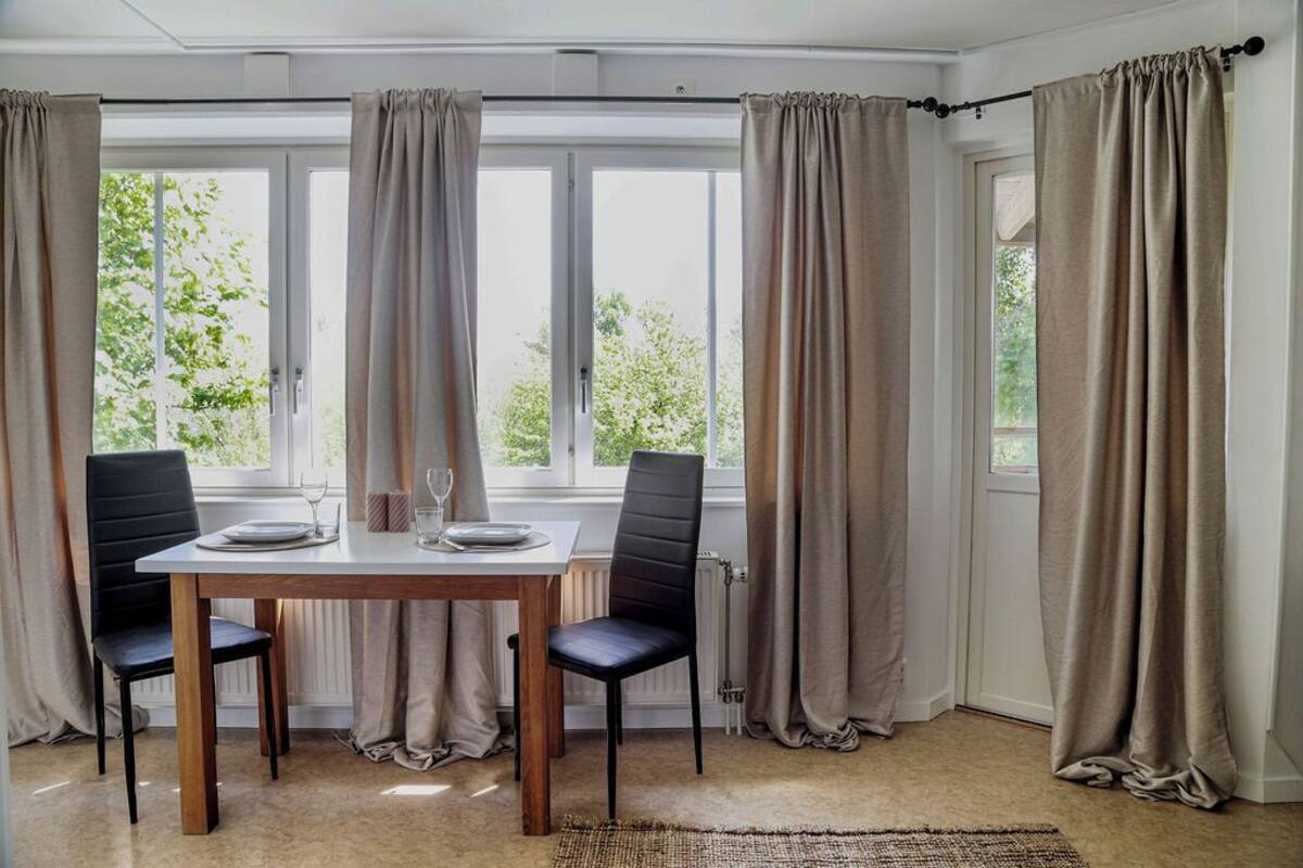 Ein Essbereich mit Tisch und Stühlen, großen Fenstern mit Vorhängen und Blick auf Grünfläche.