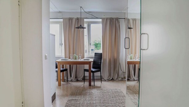A modern dining area with a wooden table set for two, beige curtains, and a textured rug. - GotlandStay Volontären (Visby)