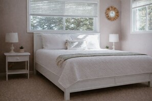 A bedroom with a bed, two bedside tables, a window with blinds, and a wall-mounted mirror.