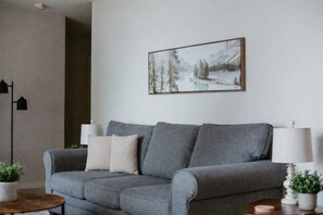 A grey sofa with two cushions, a small table with a plant, and a framed picture of a snowy landscape on the wall.