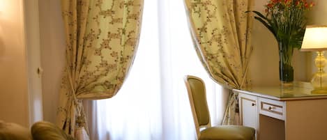 A room with a bed, a desk, and a chair. There is a window with floral curtains.