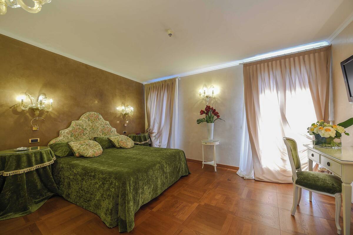 Een slaapkamer met een groen bed, een witte nachtkastje, een stoel, een bureau met een bloemenvaas, en een televisie aan de muur.