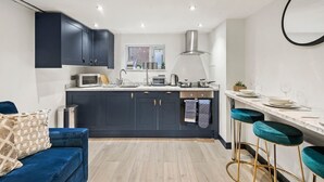 A modern kitchen with dark cabinets, a white countertop, and a window above the sink. There is a blue sofa with a patterned pillow and a small trash can.
