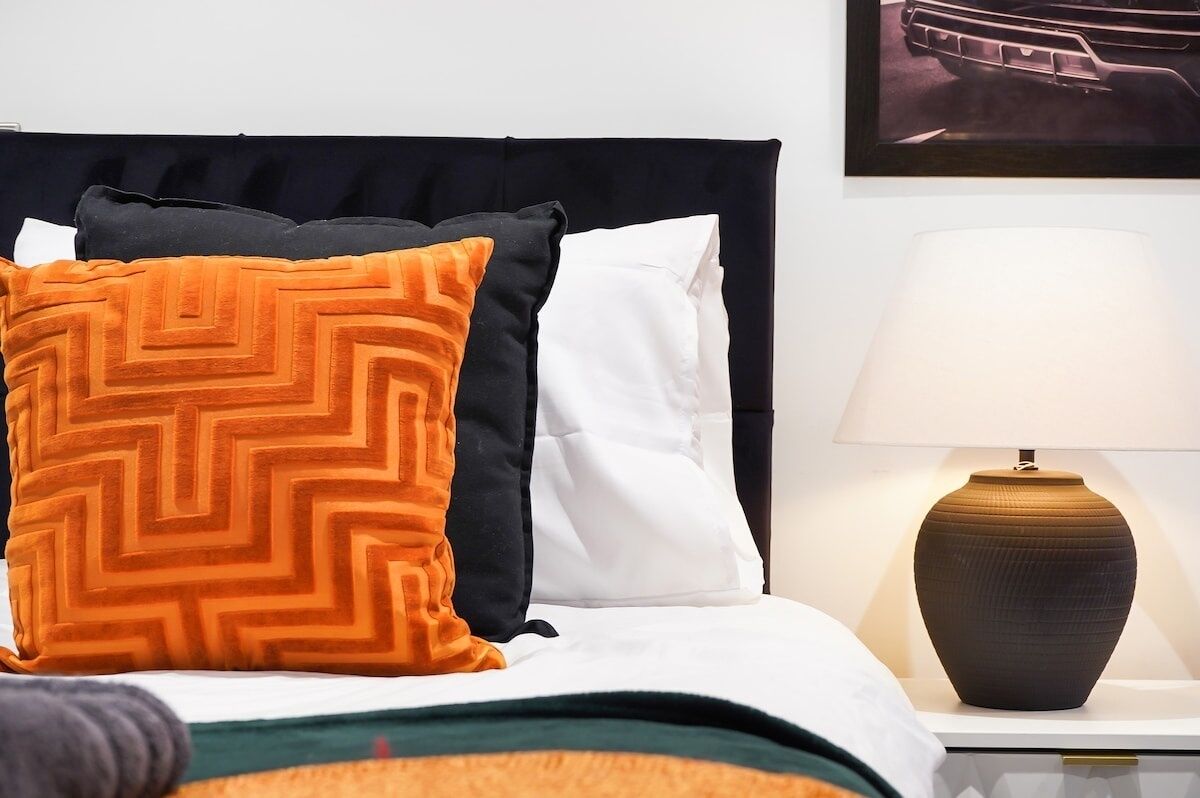 A neatly made bed with a black headboard, two pillows (one with a geometric pattern), and a white pillow. A lamp with a textured vase is on the nightstand beside the bed.