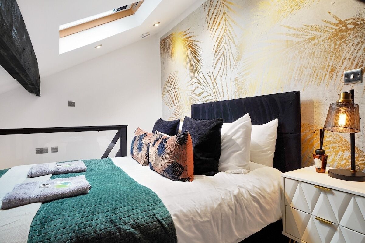 A modern bedroom with a skylight, a bed with multiple pillows, a nightstand with a lamp, and a wall with a leaf pattern.
