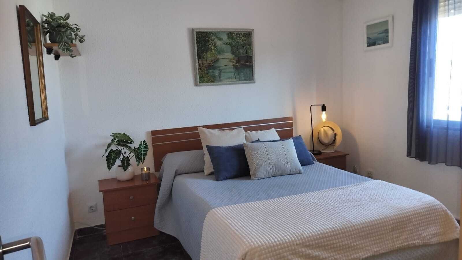 Un dormitorio ordenado con cama, mesitas de noche, una mesita auxiliar, un estante en la pared con una planta y un cuadro.