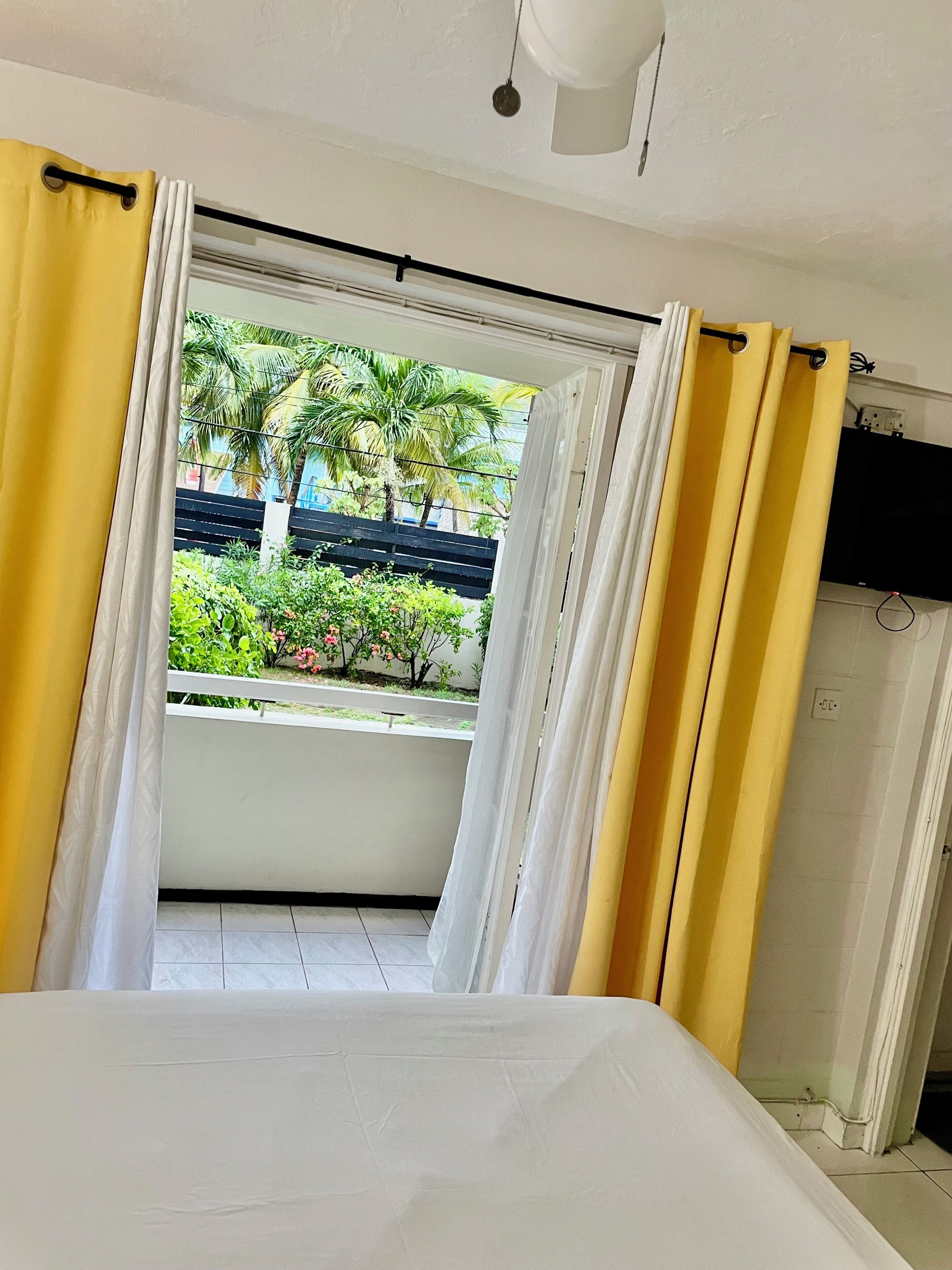 Habitació amb un llit, cortines i vista a un balcó amb plantes i una tanca.