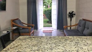 A room with a granite countertop, two wooden chairs, a small table, a standing fan, and a view of a garden through a doorway. - Ochi Beach Getaway (Unknown)