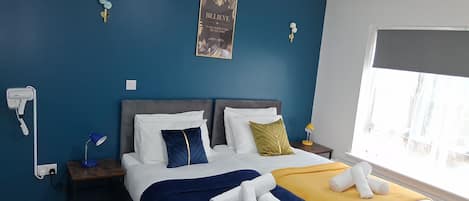 A hotel room with a bed, a bedside table with a lamp, a wall-mounted hairdryer, and a poster.