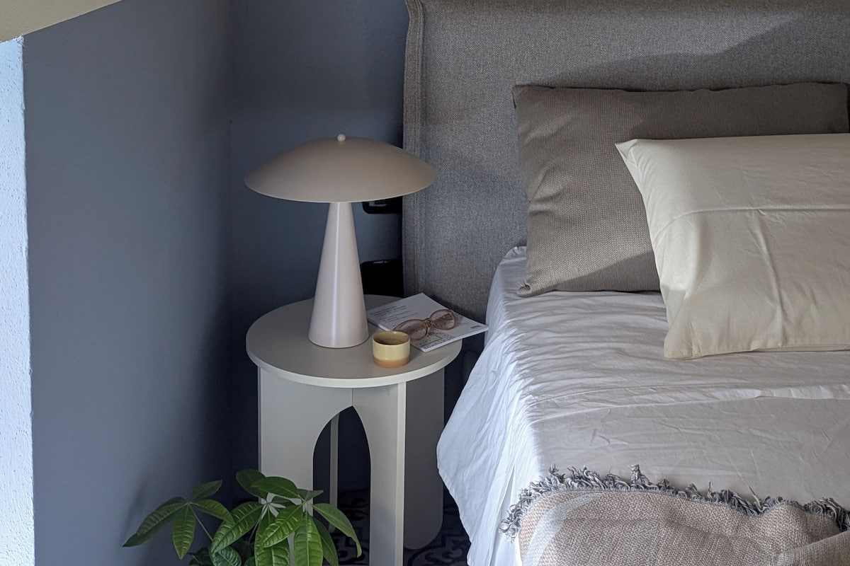 A neatly arranged bedroom with a bed, pillows, a bedside table with a lamp, and a framed picture on the wall.