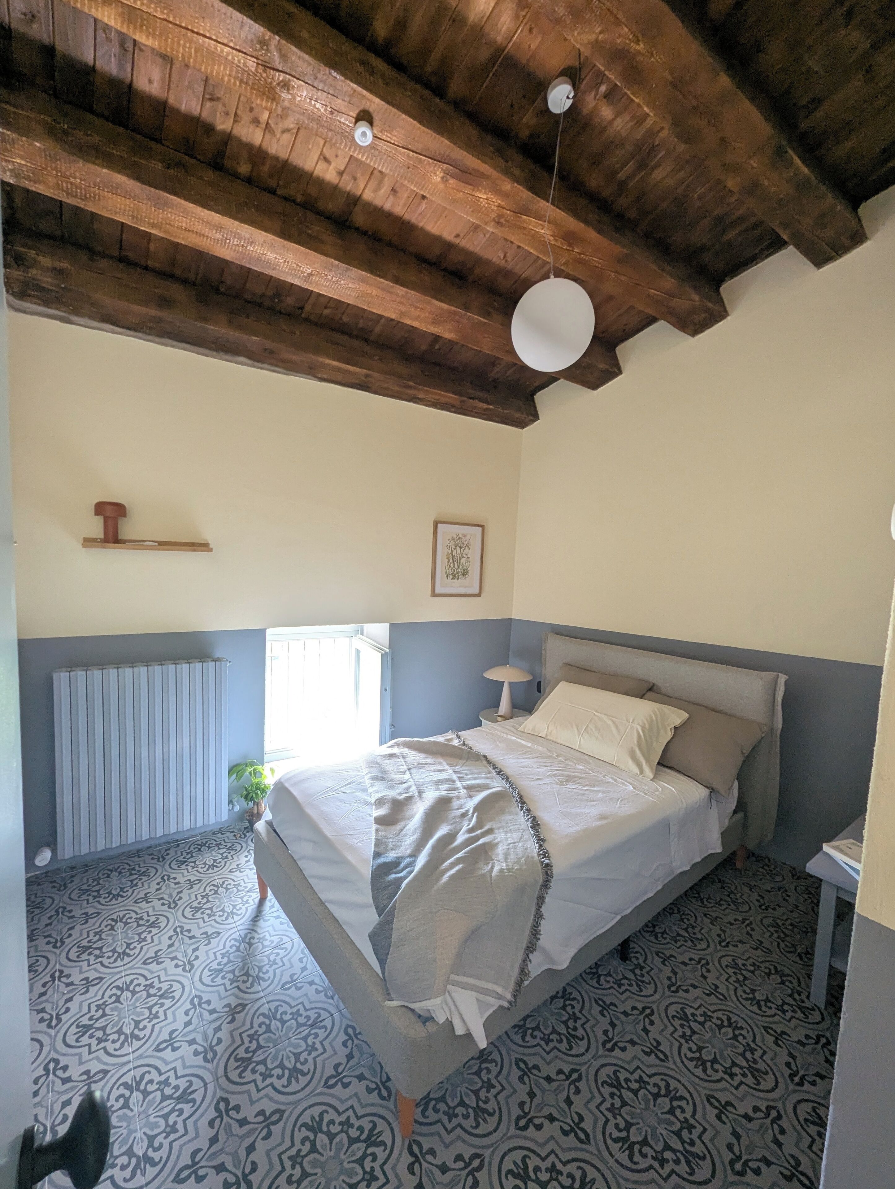 Ein Schlafzimmer mit einem Bett, einem Heizkörper, einem Fenster und einer Holzbalkendecke.