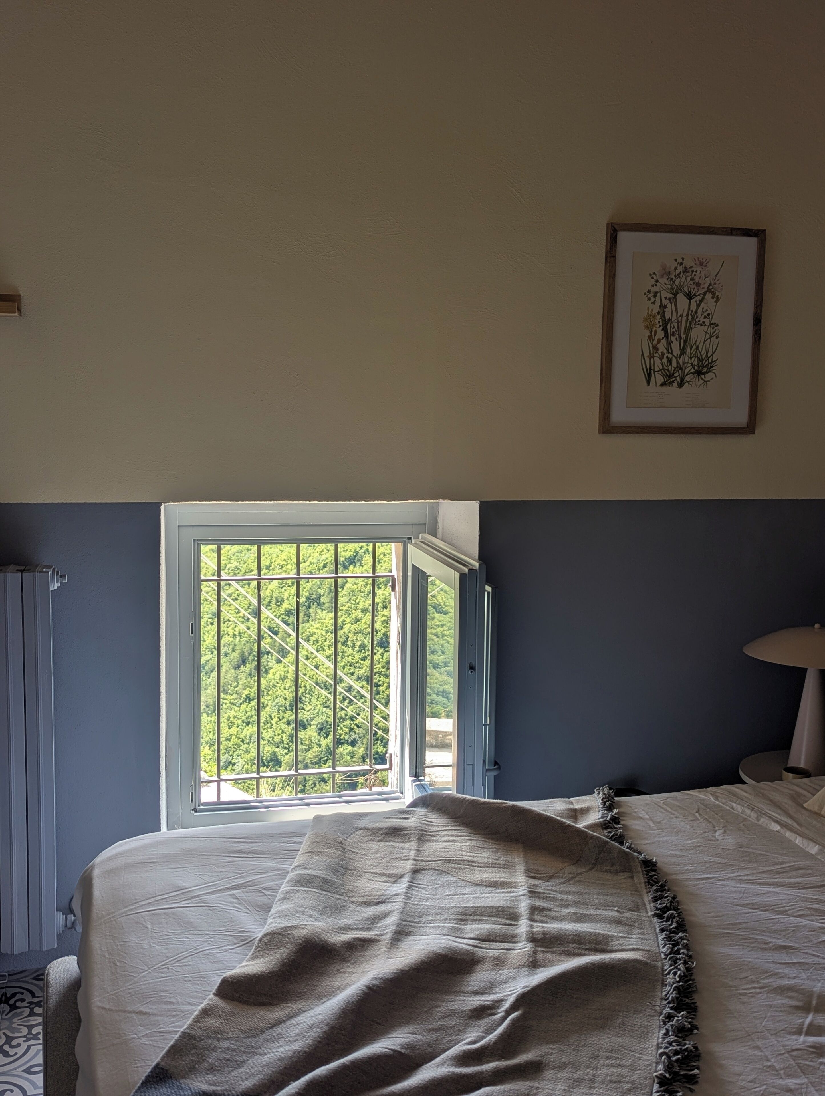 A bedroom with a bed, a window with a view of greenery, a framed botanical illustration on the wall, and a bedside lamp.