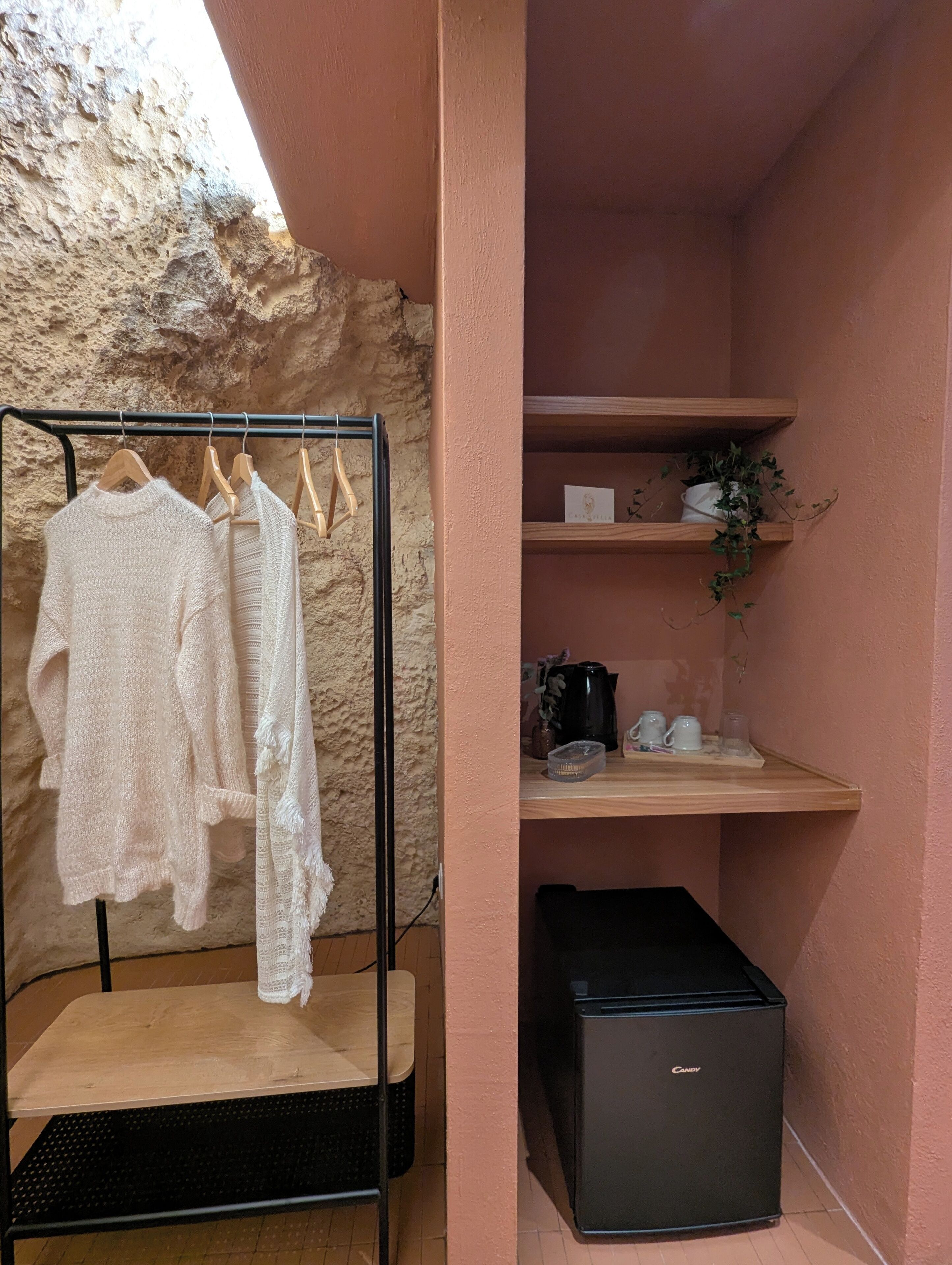 Ein Zimmer mit einem schwarzen Metallregal, das weiße Kleidungsstücke, einen schwarzen Kühlschrank und ein Holzregal mit einer Topfpflanze und Küchenutensilien beherbergt.