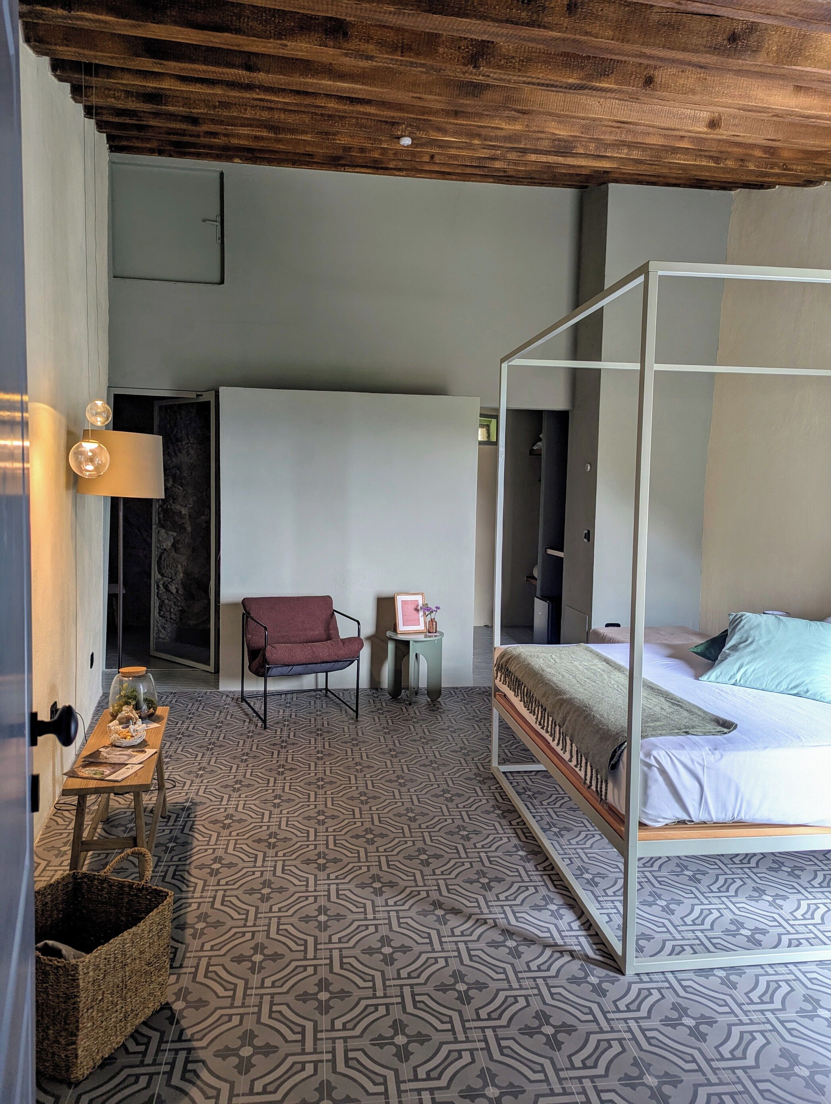 Une chambre avec un lit à baldaquin, une petite table avec une lampe, une chaise et un objet décoratif posé sur une table d’appoint.