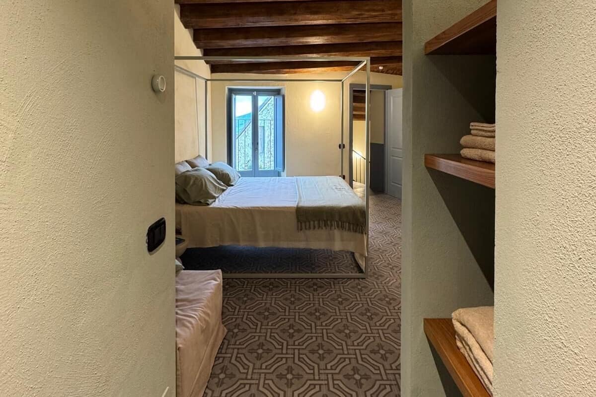 A narrow hallway leading to a bedroom with a patterned floor, wooden ceiling beams, and a bed with a window view.