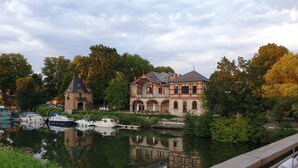 A historic building with a tower and a large arched entrance, surrounded by a calm river and lush trees.